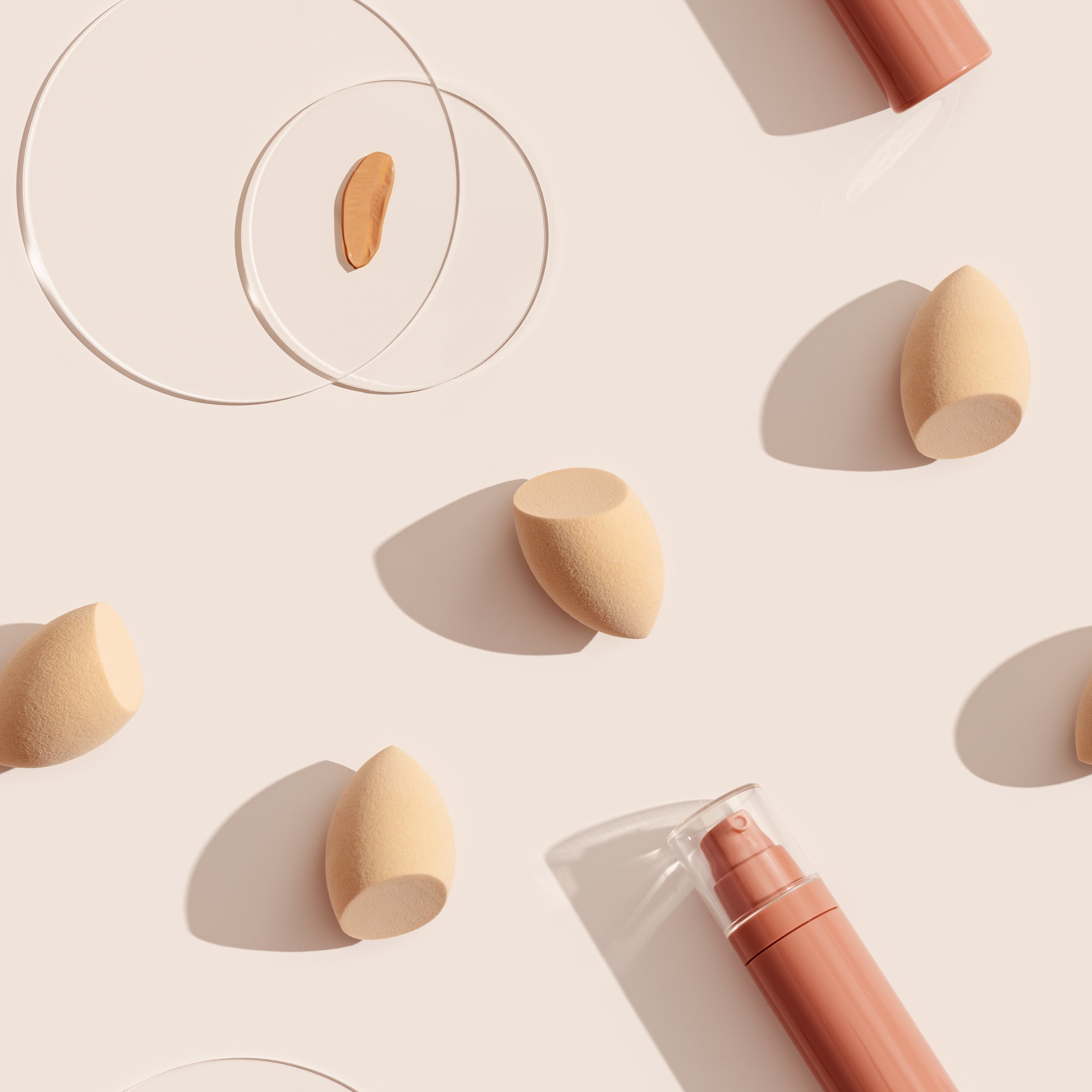 Makeup liquid foundation nude cream cosmetic bottle mockup, swat Makeup liquid foundation nude cream cosmetic bottle mockup, swatches on glass disk, beauty blender, sponges, make up puff on beige background with dark shadows. Seamless pattern, tonal cream